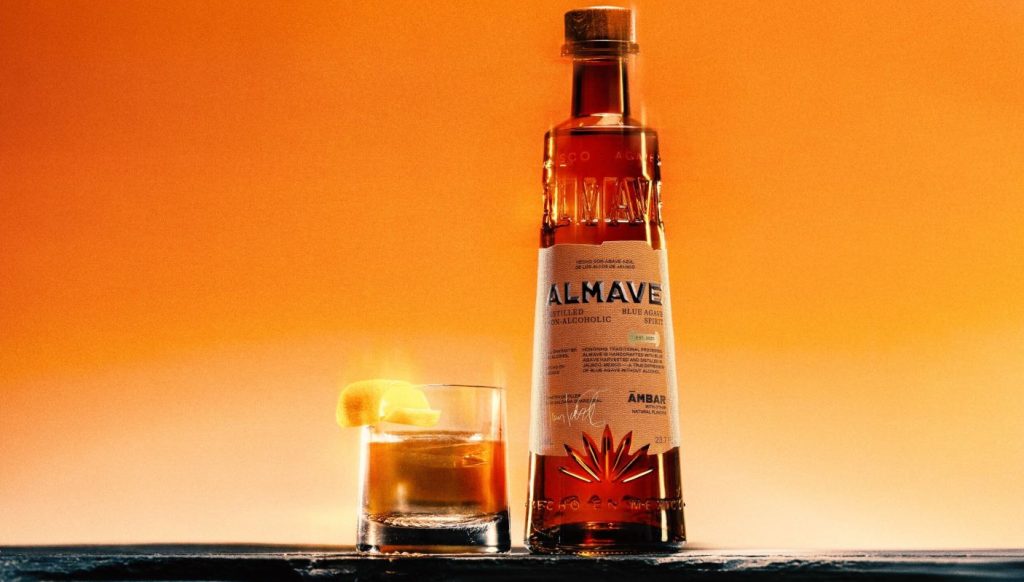 Orange bottle of Almave non-alcoholic Ambar agave-based spirit next to cocktail with citrus peel in glass on black stone countertop, orange background.