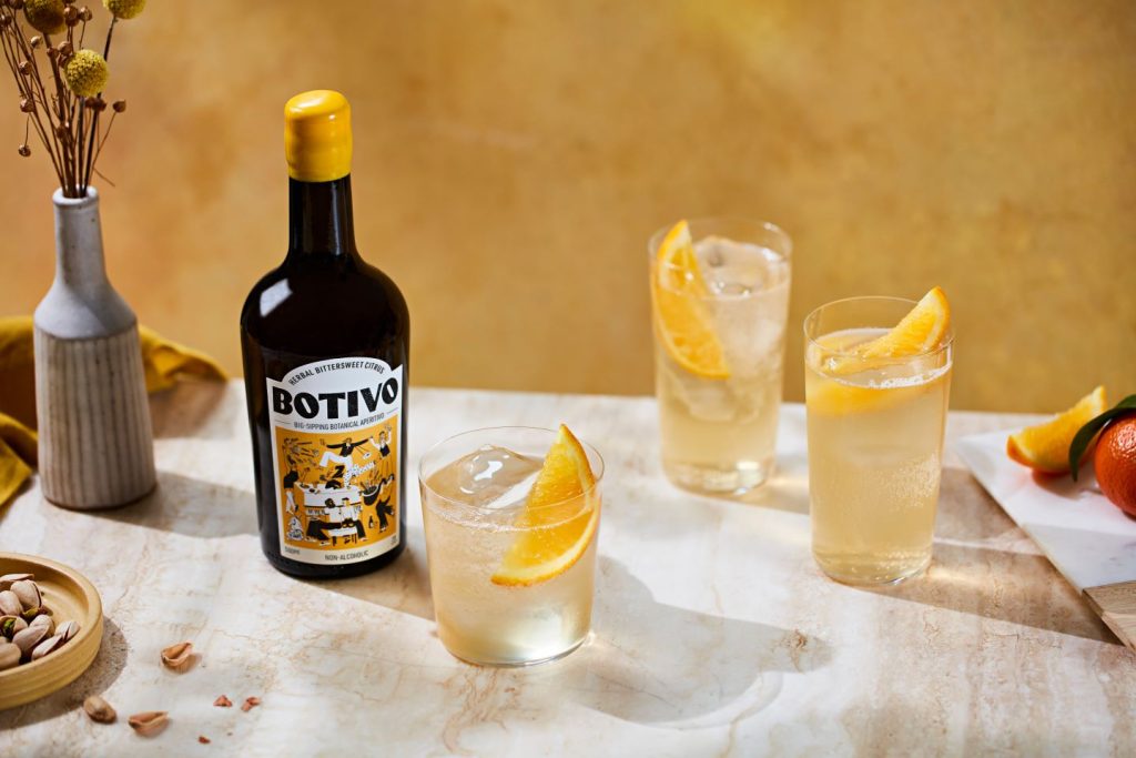 Botivo non-alcoholic aperitivo bottle with yellow top on table with glasses of aperitif and orange slices, yellow wall background