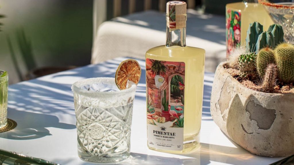 Bottle of ready-to-serve Margarita drink, Pimentae brand, next to short cocktail glass on sunlit white table. 