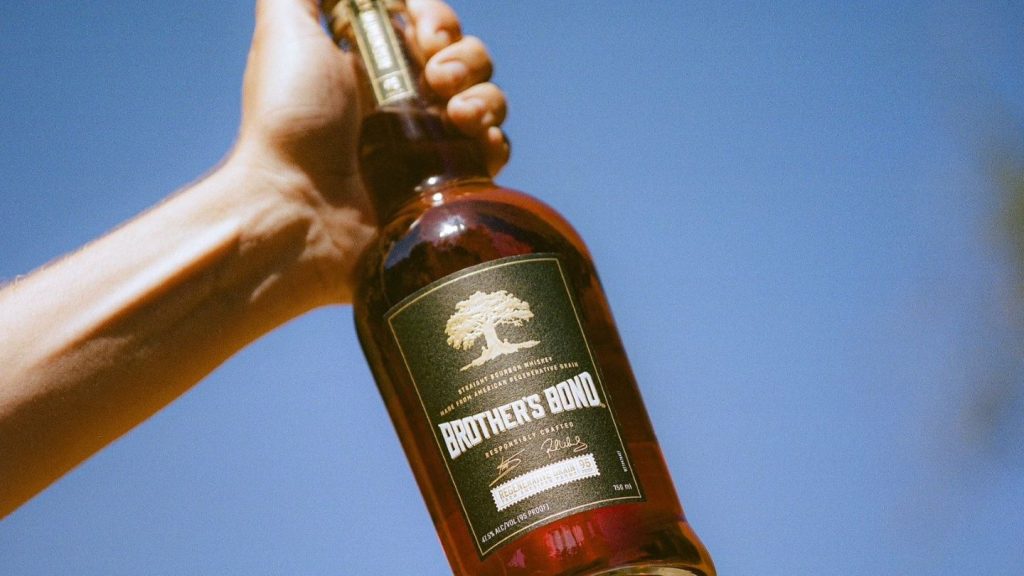 hand holding bottle of bourbon whiskey against blue sky, sun shining on bottle.