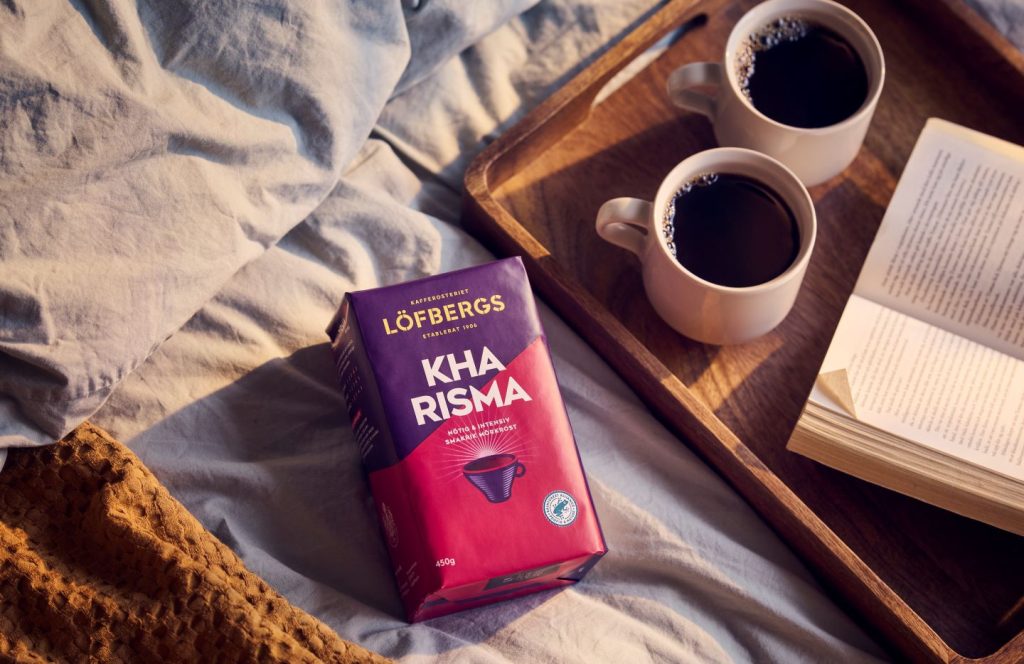 Purple, pink coffee pack on grey sheets next to wooden tray with two cups of coffee